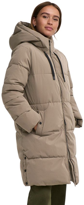 Bruine, lange winterjas met capuchon, aan de zijkant gedragen door een model met handen in de zakken.