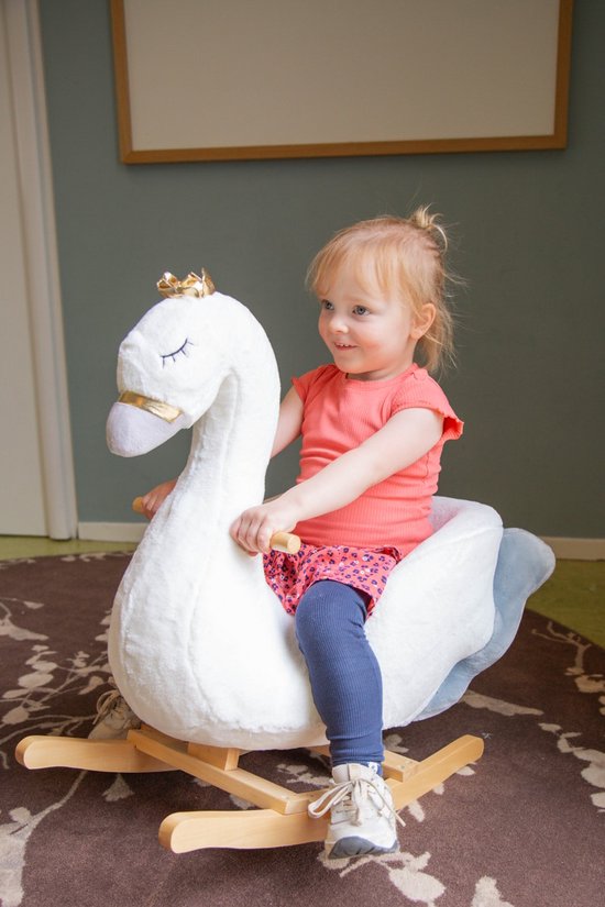 Een peuter in een oranje shirt en een roze rokje rijdt op een witte zwaan schommelstoel met gouden kroon.