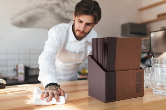 Een ober maakt een houten tafel schoon met een witte doek, naast een bruine menubox met menukaarten.