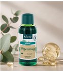 Kneipp Badolie Refreshing Eucalyptus in fles met donkergroene dop. Het etiket toont "Badolie Refreshing Eucalyptus". Naast de fles ligt een glazen object met bubbels en gele druppels, en eucalyptusblaadjes.