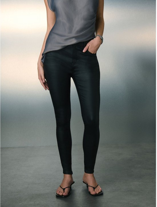 Model staat in grijze top met één schouder en zwarte skinny jeans met coating.
