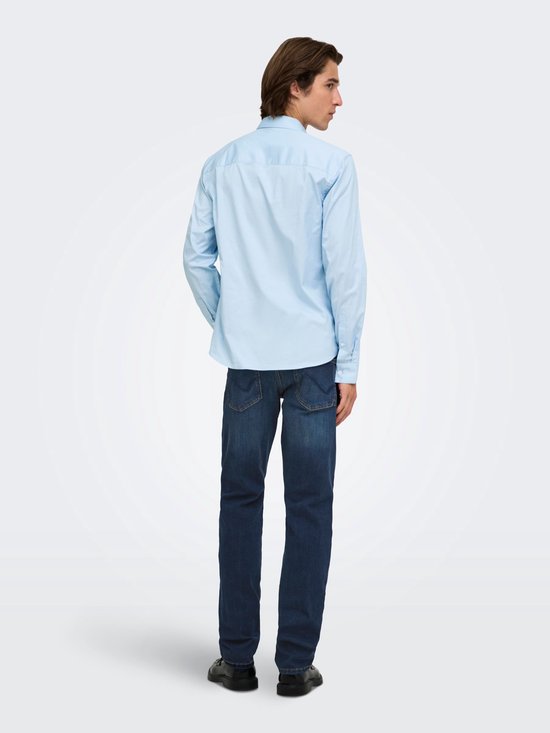 Man draagt lichtblauwe blouse met lange mouwen en donkere jeans, staand met rug naar de camera.