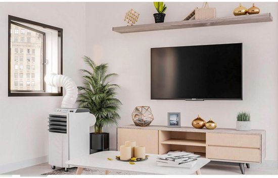 Witte mobiele airco met slang aangesloten op een open raam, naast een plant en een tv-meubel in een woonkamer.