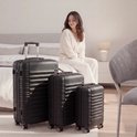 Een set van drie zwarte TRVLMORE koffers staat op een zachte vloer, met een vrouw in een witte pyjama die op een bed zit.