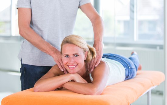 Een blonde vrouw met blauwe ogen, lachend, liggend op een oranje massagetafel, met een therapeut die haar schouders masseert.