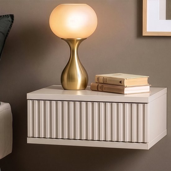 Beige hangend nachtkastje met verticale ribbels op ladefront, naast een bed met kussens en een lamp.