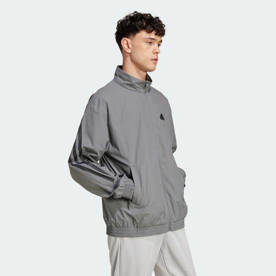 Man draagt een grijs Adidas trainingsjack, zijkant zichtbaar, met zwarte 3-strepen op de mouwen.