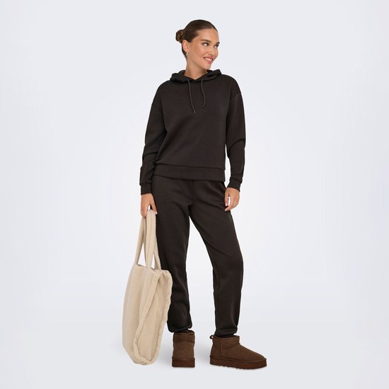 Model met donkerbruine joggingbroek en hoodie, draagt bruine laarsjes en draagt een beige teddy shopper.