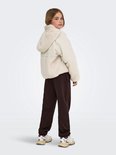 Meisje staat in een beige teddy jas, bruine joggingbroek, van achteren gezien.
