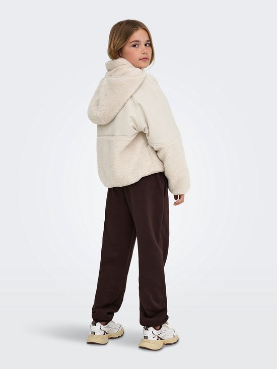 Meisje staat in een beige teddy jas, bruine joggingbroek, van achteren gezien.