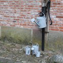 Een zinken gieter met sproeikop hangt aan een waterpomp naast een bakstenen muur. Drie kleinere gieters staan op de grond.