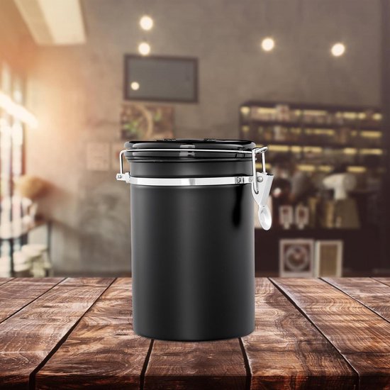 Zwarte luchtdichte koffiebus van metaal met zilverkleurige sluiting en lepel, op een houten tafel in een café.