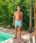 Jongen in turquoise zwembroek met een wit patroon van gebogen lijntjes, staand bij een zwembad.