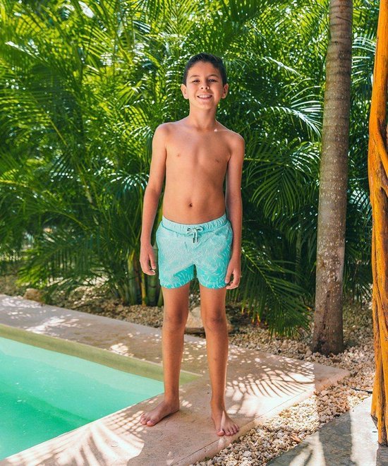 Jongen in turquoise zwembroek met een wit patroon van gebogen lijntjes, staand bij een zwembad.