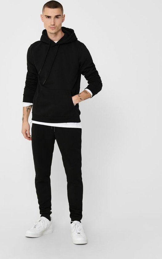 Man met tatoeages draagt een zwarte hoodie over een wit shirt met joggingbroek.