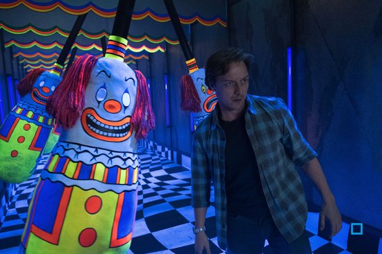 Man staat in een gang met kleurrijke clown-figuren en een zwart-wit geblokte vloer.