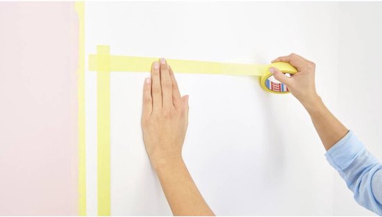 Vrouw plakt gele Tesa tape op een witte muur langs een roze muur.