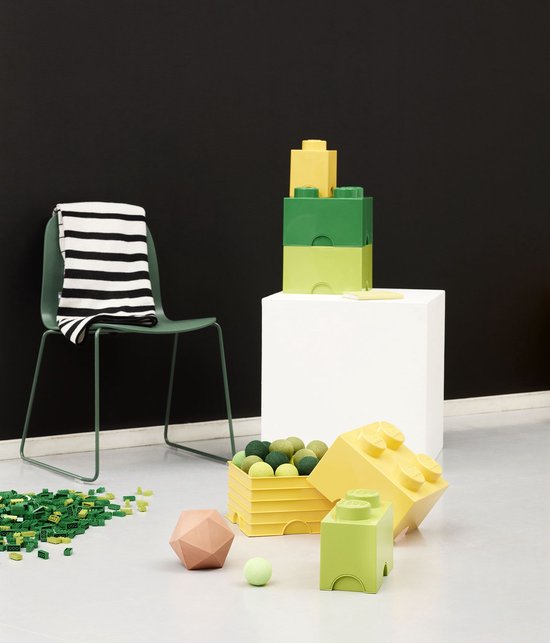 Groene en gele Lego opbergdozen gestapeld op een witte kubus, naast een groene stoel met gestreepte deken.