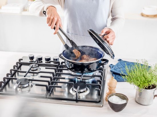 Een persoon gebruikt een tang om gebakken burgers uit een blauw-zwarte braadpan te halen. De pan staat op een gasfornuis.