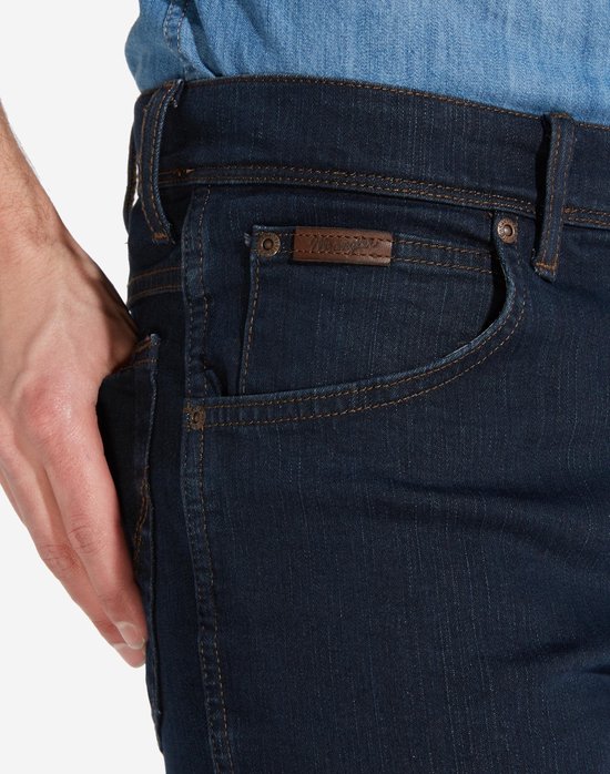 Close-up van de voorzak van een donkerblauwe Wrangler jeans met kleine merkpatch en koperkleurige klinknagels.
