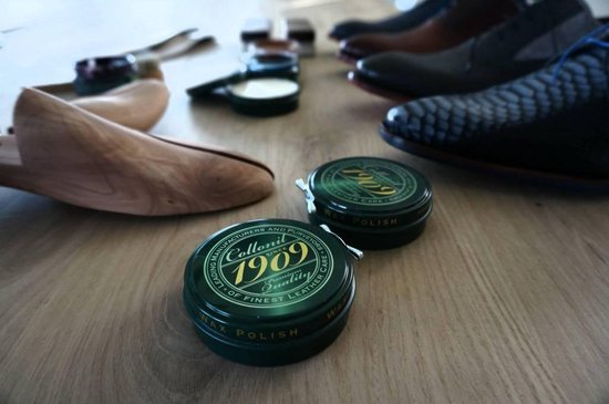 Twee potjes Collonil 1909 wax polish op een houten ondergrond, met schoenen en leesten op de achtergrond.