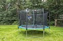 Een kind springt op een groene trampoline met veiligheidsnet, in een tuin met bomen.