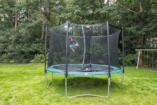 Een kind springt op een groene trampoline met veiligheidsnet, in een tuin met bomen.