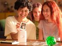 Een hand houdt een Instax foto omhoog, met drie lachende personen op de foto. Op tafel staat een witte Instax Mini Link 2 printer.