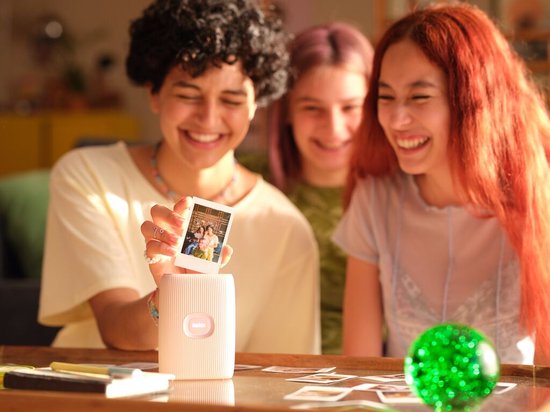Een hand houdt een Instax foto omhoog, met drie lachende personen op de foto. Op tafel staat een witte Instax Mini Link 2 printer.