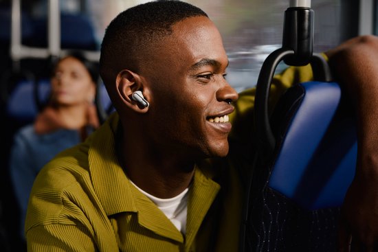 Man met een groen vest draagt een zwart JBL oordopje terwijl hij in de bus zit.