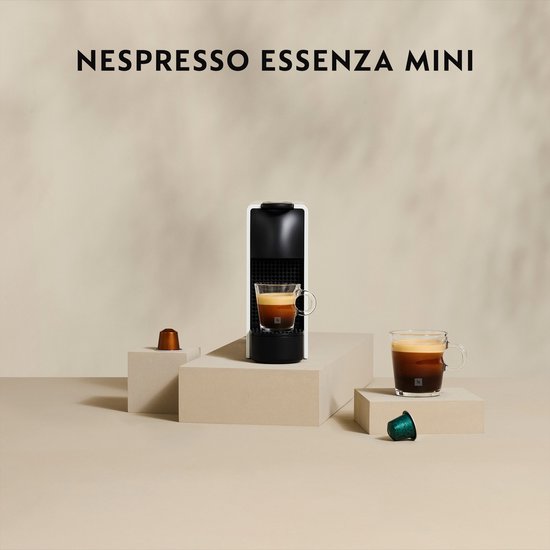 Machine à café Nespresso Essenza Mini blanche, à côté de deux capsules de café et avec deux tasses d'expresso.
