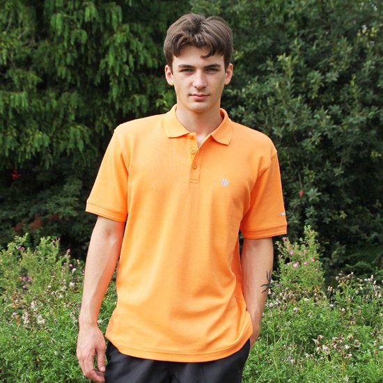Model in een feloranje polo met korte mouwen en een klein logo op de borst, staand in de natuur.