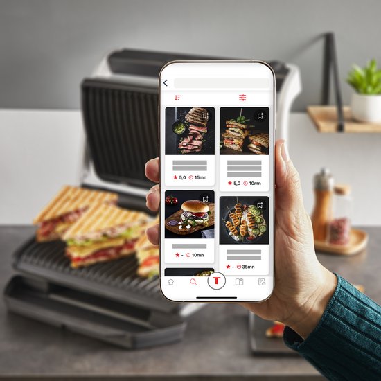 Hand houdt smartphone met recepten-app. Op het scherm staan foto's van gegrilde gerechten en de contactgrill staat op de achtergrond.