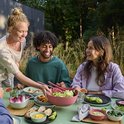 Groep vrienden geniet van een diner in de tuin. Een vrouw serveert een roze schaal met salade. Servies in roze en groen.