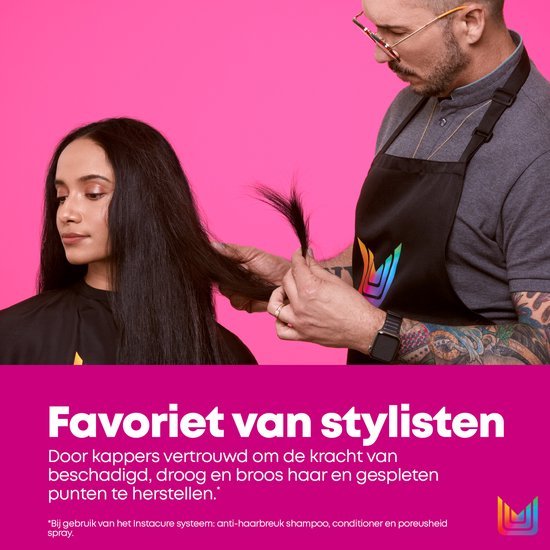 Coiffeur avec lunettes et tatouages coupe les cheveux longs et foncés d'une femme brune. Fond rose.