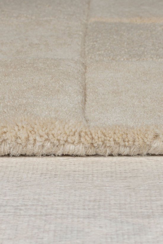 Close-up van de rand van een beige, hoogpolig vloerkleed met blokpatroon.