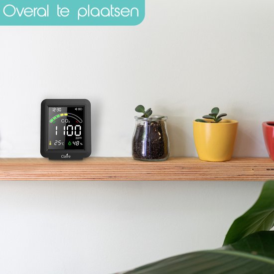 Claire Airbox CO2 meter op een houten plank, met planten. De meter toont 1100 ppm CO2, 25°C, 48% luchtvochtigheid.