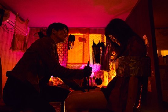 Man in donker shirt met mes, boven een vrouw in rood, in een kamer met felroze verlichting.