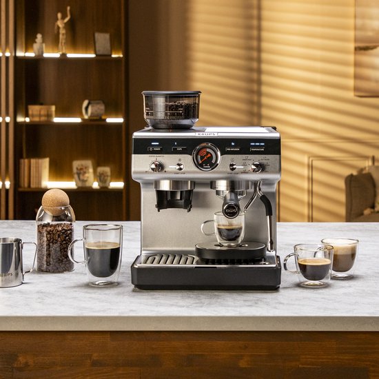 Zilverkleurige Krups espressomachine met ingebouwde grinder op een aanrecht met koffiebonen en kopjes.