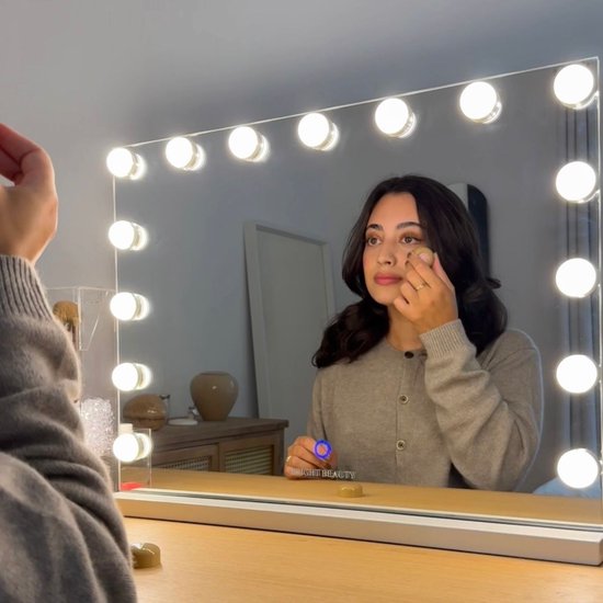 Vrouw met bruin haar en grijze cardigan gebruikt make-up in spiegel.