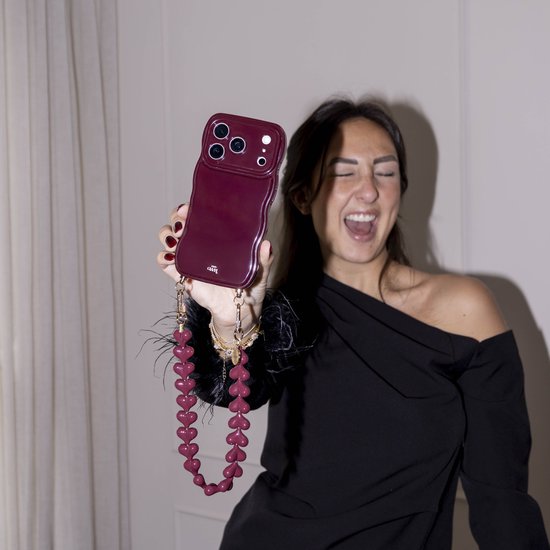 Bordeauxrode, golvende iPhone-hoes met bijpassend koord van hartjes. Vastgehouden door een vrouw met zwarte veren op haar arm.