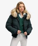Model in een donkergroene, gewatteerde bomberjack met bruine bontkraag. Draagt een gestreepte blouse en groen vest.