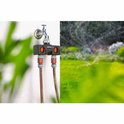 Gardena waterverdeler met kraan en twee slangen. Rechts een tuin met sproeiers. Links de muur waar hij aan vastzit.