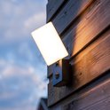 Wandlamp met een breed wit lichtpaneel en sensorbehuizing op een houten schuur.