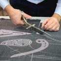 Een naai-liniaal met roze markeringen ligt op donkergrijze stof, een schaar snijdt de stof.