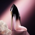 Roze en zwarte pump-vormige parfumfles met gouden hak, naast een witte bloem.