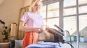 Vrouw met roze shirt en oranje broek, vult een doorzichtige watertank van een zwarte stoomgenerator. Een gestreken paars shirt ligt klaar.