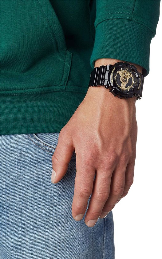 Man met groene trui en spijkerbroek draagt een zwart G-Shock horloge met gouden wijzerplaat.