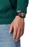 Man met groene trui en spijkerbroek draagt een zwart G-Shock horloge met gouden wijzerplaat.