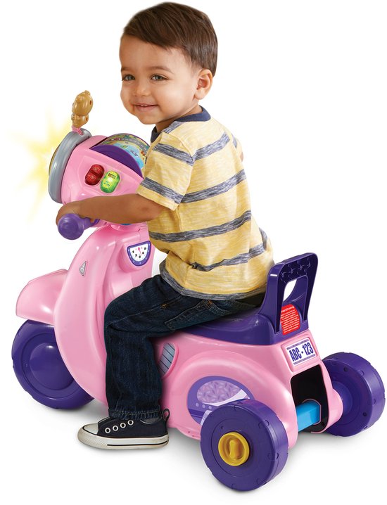 Jongen zit op roze VTech driewieler loopscooter met paarse details en knipperende lampjes.